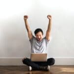 Photo of Man in Gray T-shirt and Black Pants Sitting on Wooden Floor and Working on His Laptop Celebrating with His Hands Raised
