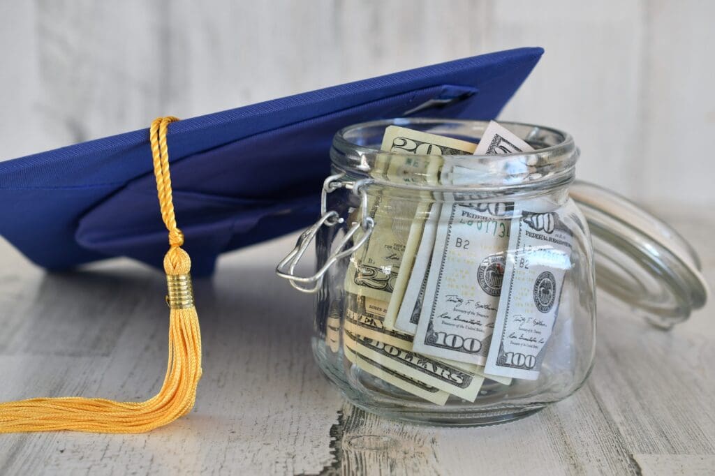 Graduation cap mortarboard with tassel propped on a jar of money cash, concept school loans debt