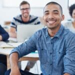 Portrait of a young office worker sitting at a table with colleagues in the background
