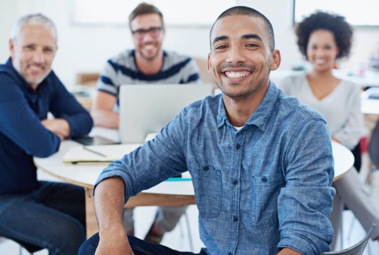 Portrait of a young office worker sitting at a table with colleagues in the background