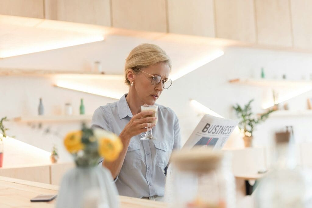 mature businesswoman in eyeglasses reading newspaper and drinking coffee in cafe reading about small business news