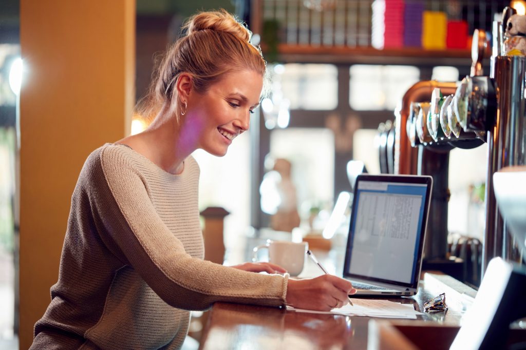 Female Owner Or Manager Of a fast-paced Bar Working On Laptop On Counter