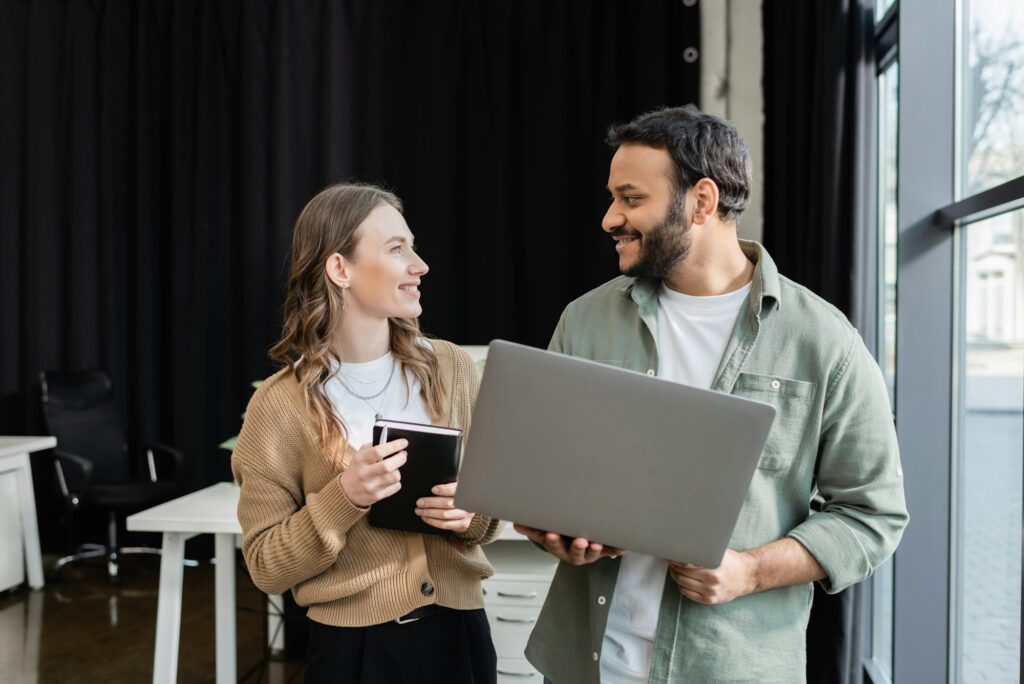 happy businessman with laptop explaining task to female coworker with notebook, mentorship