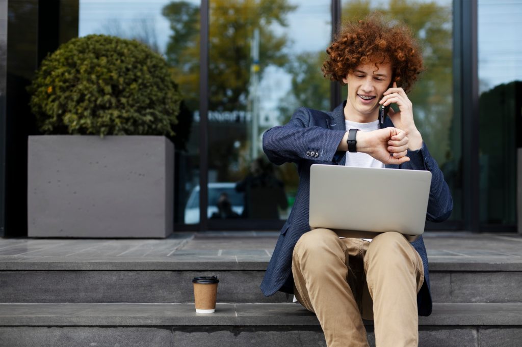 Successful handsome programmer using laptop, checking the clock, talking on cellphone