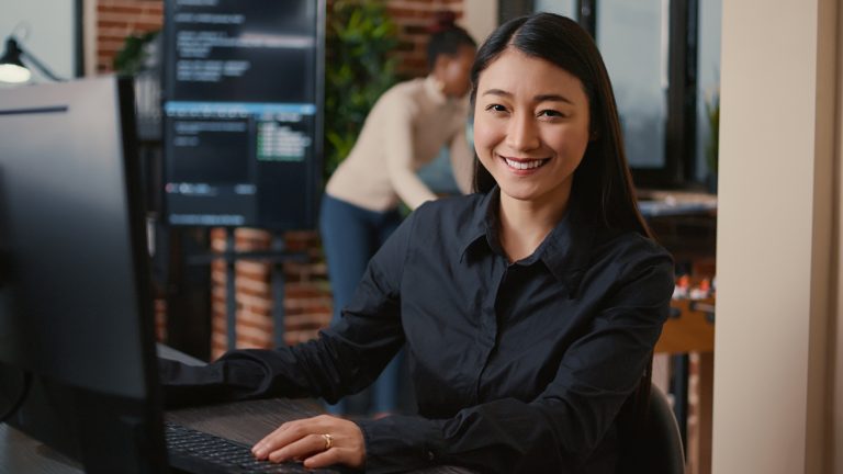 Asian software developer smiling and resuming work typing code on computer keyboard