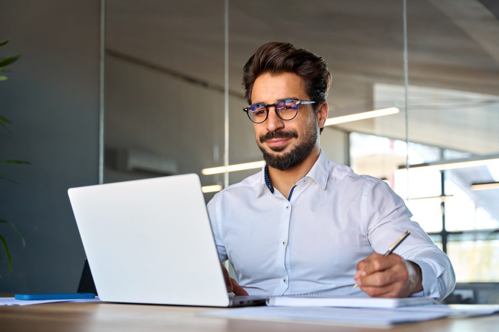 Busy smiling young Latin business man working in office using laptop writing.