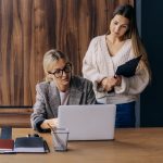Two positive young women working and discussing together at office table coworking.