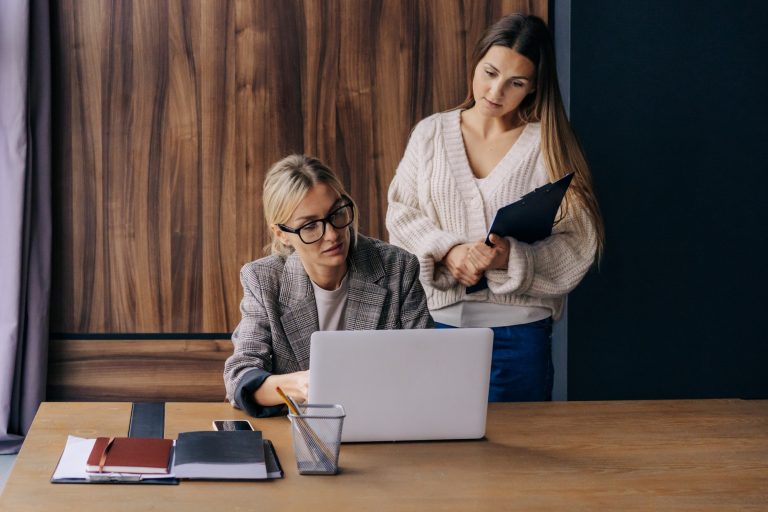 Two positive young women working and discussing together at office table coworking.