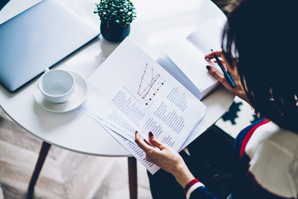 Intelligent female expert analyzing paper report with graph charts sitting at table with laptop