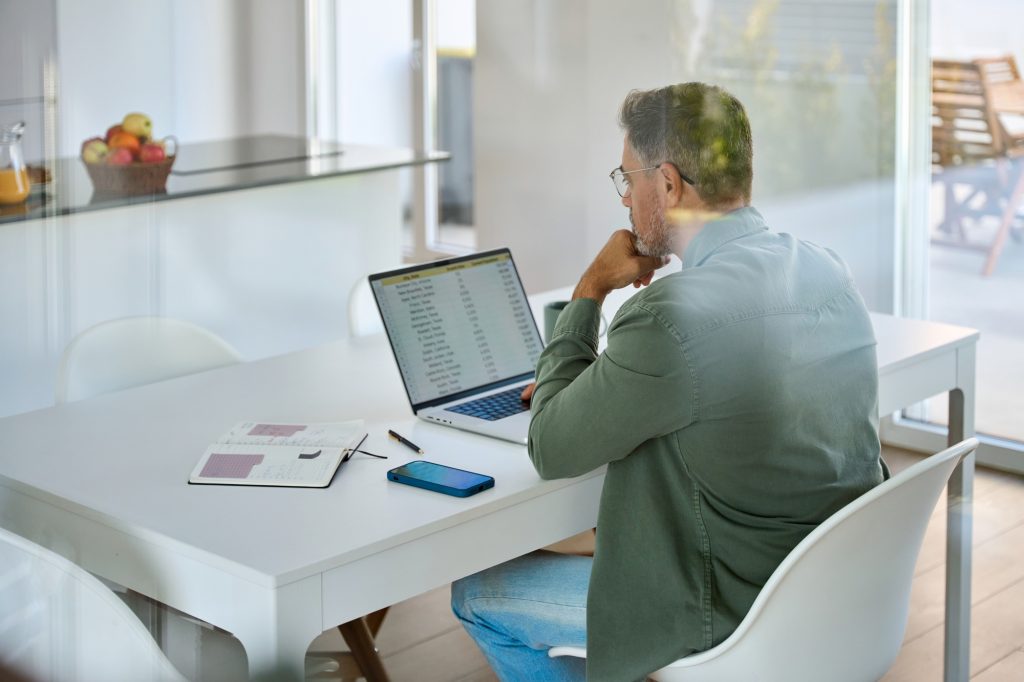 Mature man looking at computer sitting at kitchen table hybrid working at home.