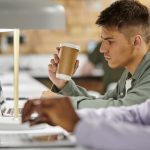 young man holding coffee to go and using laptop near african american colleague, startup, gen z