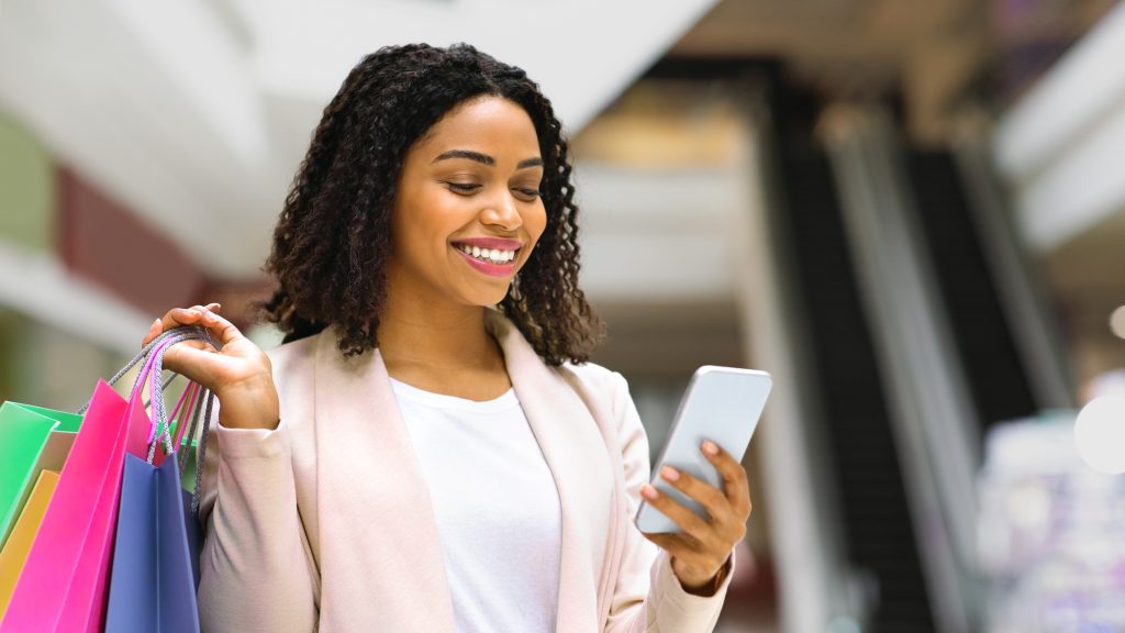 Discount App. Smiling Woman With Shopping Bags Using Smartphone In Mall